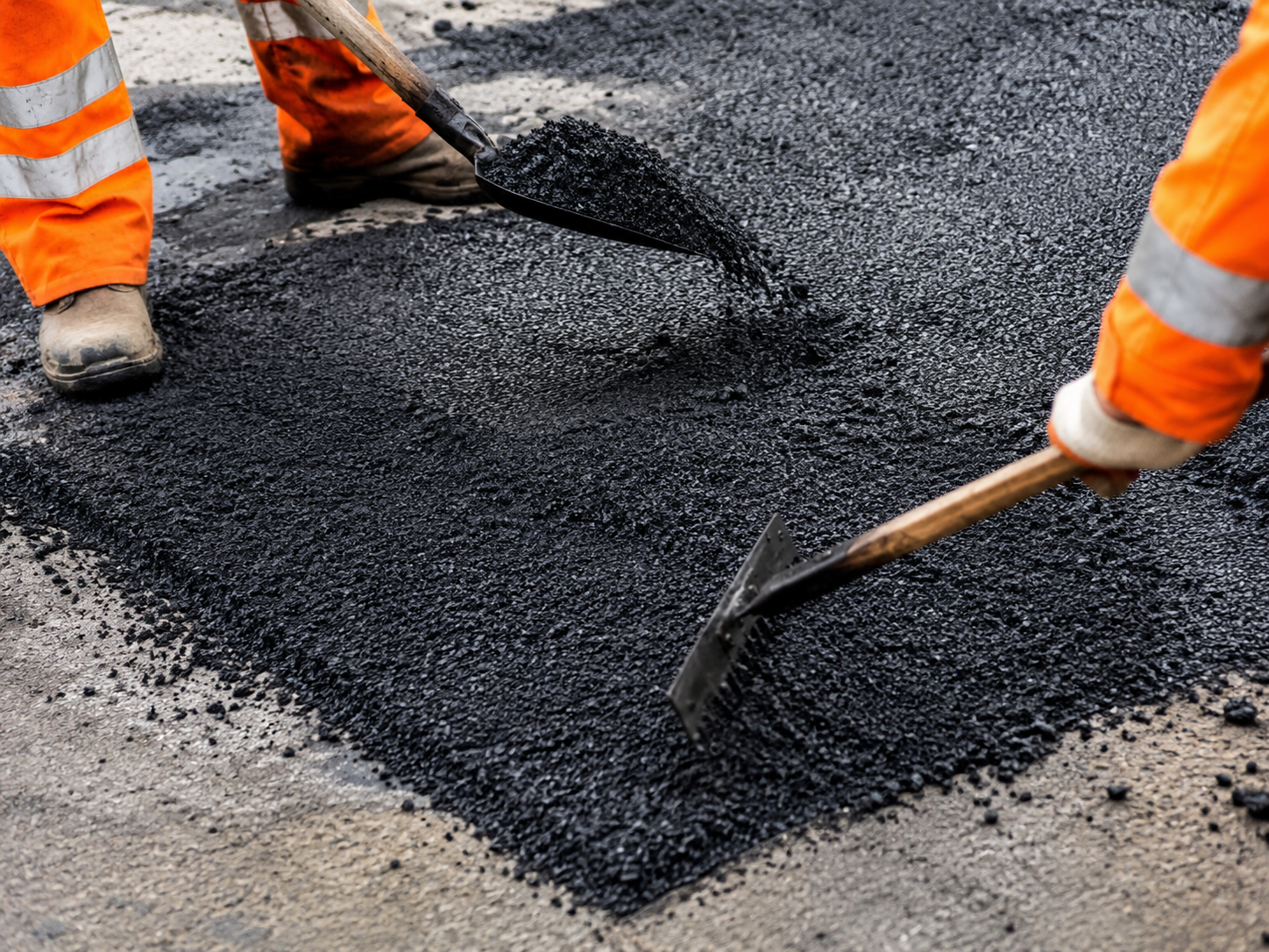 Two road crew in orange hi-vis shovelling and raking fresh hot asphalt mix onto a road repair.