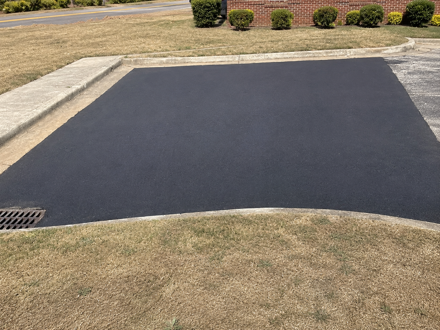 Completed asphalt resurfacing patch in front of a brick building, neatly cut into the kerb and grass border.