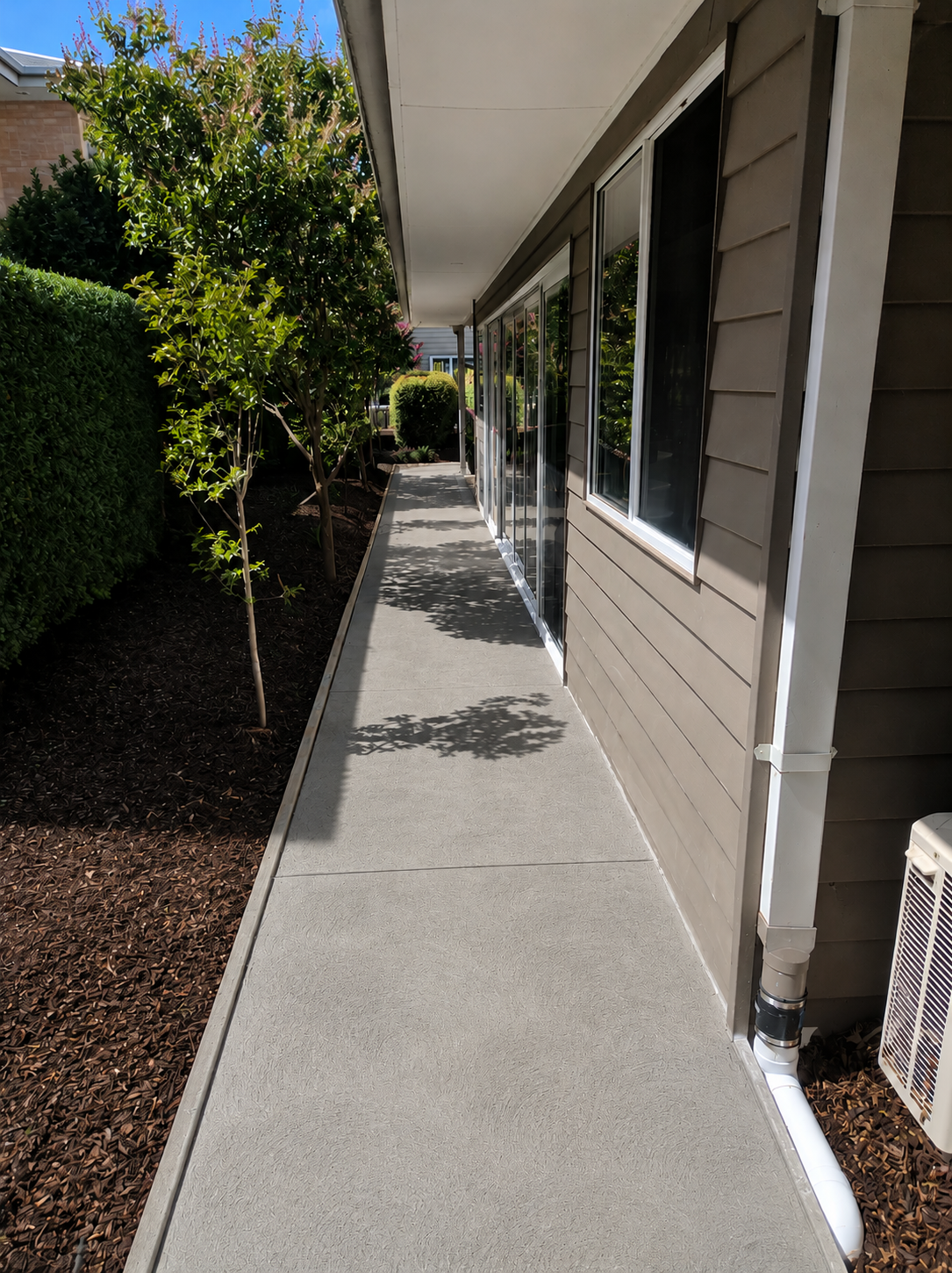 Newly poured swirl-finish concrete pathway alongside a weatherboard home, bordered by garden beds and mulch.