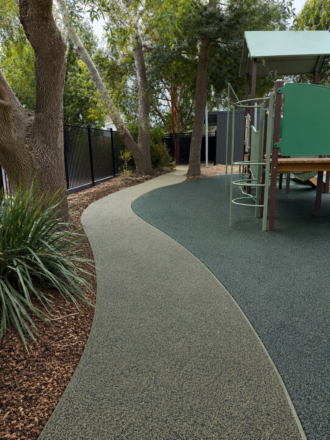 Curved permeable surface pathway next to a playground, finished in green wetpour and bordered by trees and garden beds.